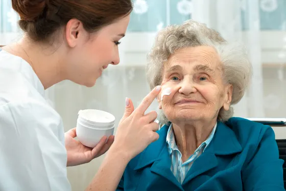 Opiekunka delikatnie nakłada krem na twarz uśmiechniętej seniorki
